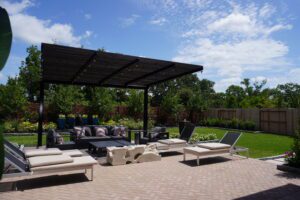 Outdoor patio area with a pergola, contemporary furniture including sofas and loungers, set against a background of manicured lawn and shrubbery under a clear sky, crafted by Texas builders.