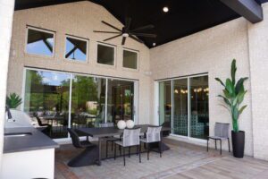 A modern outdoor patio area with a dining set, ceiling fan, potted plants, and large windows reflecting the garden, crafted by Texas builders.