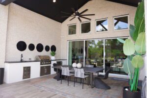 Modern outdoor kitchen and dining area constructed by Texas builders with built-in appliances and a large sliding glass door, accented by tropical plants and geometric decor.