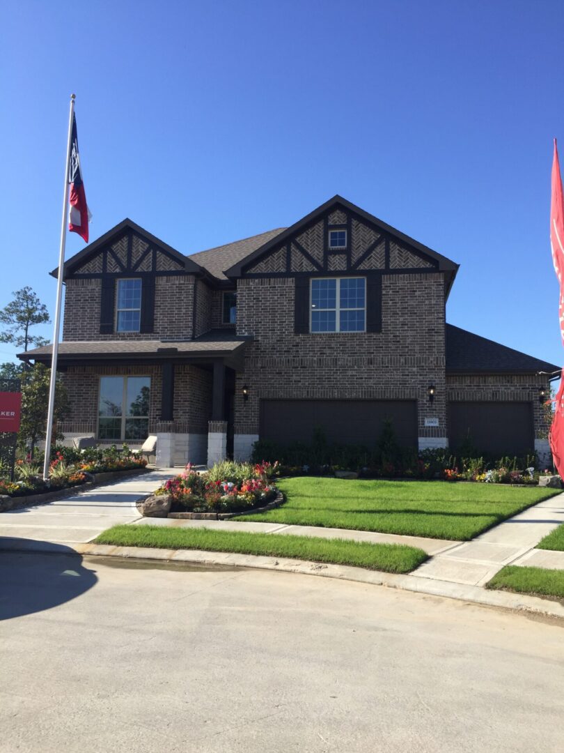 A two-story brick house with a landscaped front yard, under a clear blue sky, flanked by two waving Texas flags.