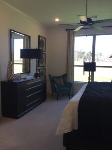 A well-lit bedroom featuring a large bed, a dresser with a mirror, a blue armchair, and large windows overlooking a grassy area constructed by Texas builders.