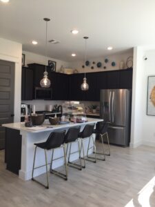 A modern kitchen designed by Texas builders with dark cabinetry, a central island with bar stools, stainless steel appliances, pendant lights, and decorative items on top shelves.