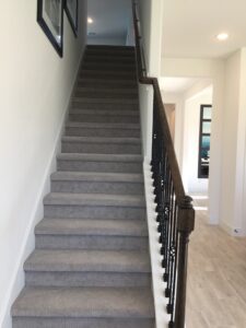 A residential staircase with gray carpeting and a dark wooden handrail, constructed by Texas builders, leading up to a well-lit upper floor.
