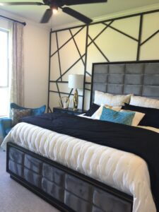 Modern bedroom featuring a large bed with a tufted gray headboard, geometric black wall design by Texas builders, white bedding, a side table with a lamp, and sheer curtains.