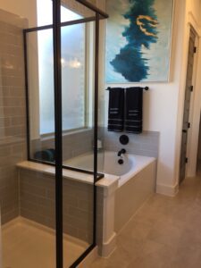 A modern bathroom featuring a walk-in shower with glass doors, a built-in bathtub, beige tiles, and a blue abstract painting on the wall, designed by Texas builders.