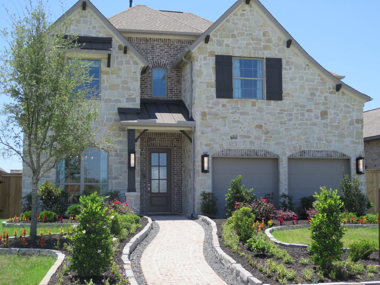 A suburban two-story house with a stone facade, dual garage doors, and a curved stone pathway lined with colorful flower beds, designed by Texas builders.