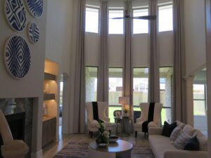 A modern living room with high ceilings, large windows, white armchairs, and a fireplace, decorated in a neutral color palette with blue accents by Texas builders.