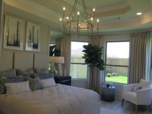 Elegant bedroom featuring a large bed with neutral bedding, abstract artwork above, a luxurious chandelier crafted by Texas builders, and a seating area near a large window with garden view.
