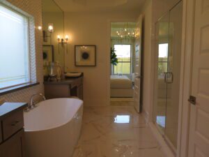 Luxurious bathroom with a freestanding bathtub, glass-walled shower, and polished floor, crafted by Texas builders, leading to a bedroom visible through an open door.