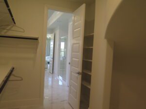 Open white door leading from a closet with shelves into a brightly lit bathroom with tiled flooring, constructed by Texas builders.