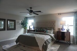 A neat bedroom with a large tufted headboard, beige bedding, side tables, lamps, and a ceiling fan designed by Texas builders, accented by a green plant and framed wall art.