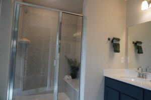 Modern bathroom designed by Texas builders, featuring a glass-door shower with geometric tile pattern, navy blue vanity, and mirror reflecting hanging green towels.