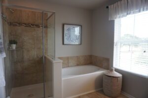 A modern bathroom featuring a glass shower stall, a large bathtub, and beige tiled walls. A window with blinds installed by Texas builders provides natural light.