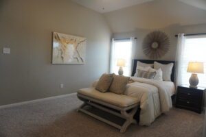 A contemporary bedroom designed by Texas builders featuring a neatly made bed with beige and white bedding, flanked by two lamps on nightstands, and decorated with a wall-mounted art piece and a sunburst mirror