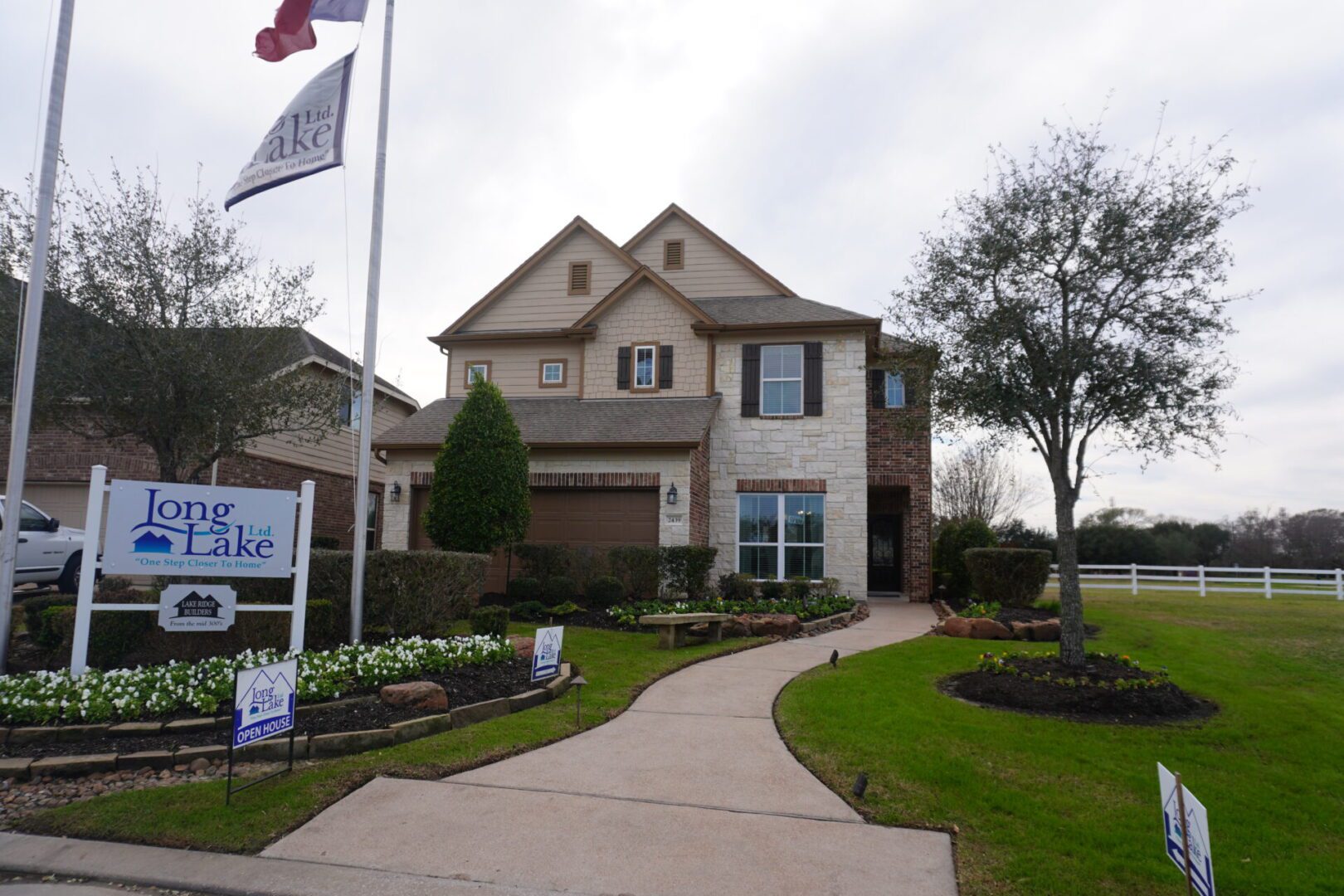 A two-story house with a brick and siding exterior, lawn with white flowers, under a cloudy sky, displaying multiple "Texas builders" real estate signs.