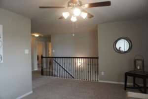Interior of a well-lit room featuring gray walls, carpeted floor, a ceiling fan with lights, and a black railing with a small mirror on the wall, completed by Texas builders.