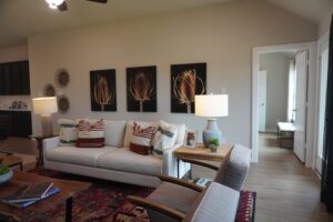 A cozy living room with a beige sofa, decorative pillows, a patterned rug, two armchairs, a lamp, and three wall art pieces showing Texas wheat motifs.