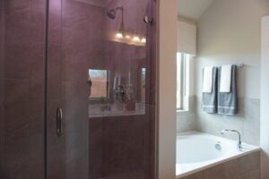 Modern bathroom with a glass shower and a large bathtub, featuring purple tiles and illuminated by a wall-mounted light fixture. Towels hang neatly beside the bathtub, expertly installed by Texas builders.