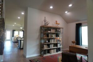 Modern home interior featuring a wooden bookshelf with decorative items, a hallway leading to an open kitchen, and a wooden figurine on top of the shelf, designed by Texas builders.