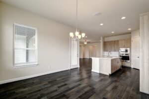 Empty modern kitchen and living space with dark hardwood floors, white cabinets, and a hanging chandelier, crafted by Texas builders.