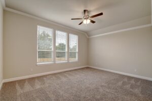 Empty room with beige walls, carpeted floor, and a ceiling fan, featuring two windows with blinds overlooking an outdoor area constructed by Texas builders.