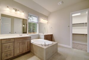 Modern bathroom interior designed by Texas builders, featuring dual sinks, a large mirror, bathtub, and a separate walk-in closet. Neutral colors and tiled flooring.
