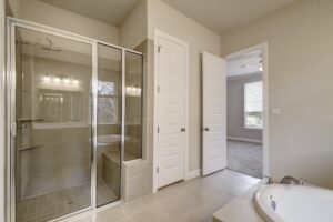 Modern bathroom designed by Texas builders, with glass shower enclosure, white doors, and a bathtub under a window, featuring neutral tones.