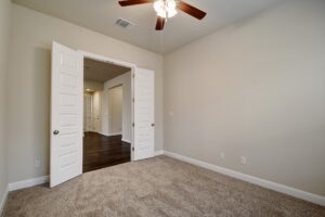 Empty room with beige walls and carpet, featuring an open door leading to another room and a ceiling fan, constructed by Texas builders.