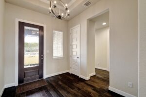 Interior of a modern home entryway crafted by Texas builders, featuring light textured walls, dark wooden floors, a stylish chandelier, and doors leading to different rooms.