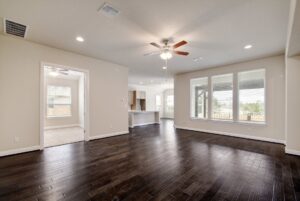 Empty living room with dark hardwood floors, ceiling fan, and large windows installed by Texas builders providing natural light, leading to adjacent rooms.