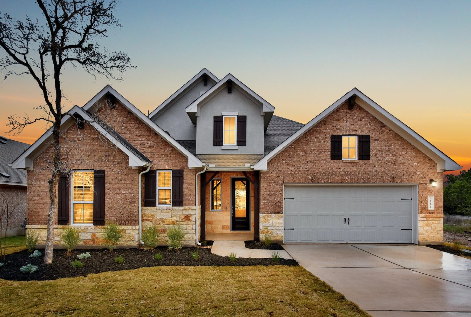 A modern brick house with gray accents and a double garage, illuminated by lights at twilight with a vibrant sunset in the background, expertly crafted by Texas builders.