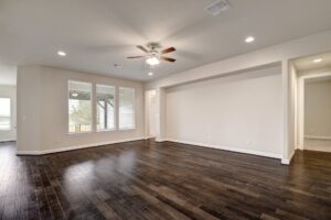 Empty living room with dark hardwood flooring, white walls, and a ceiling fan, featuring large windows and an open doorway. Built by Texas builders.
